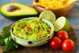 A delicious traditional bowl of guacamole next to fresh ingredients on a table with tortilla chips and salsa tasty