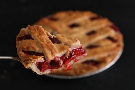 A high angle close up horizontal photograph of a slice of homemade cherry pie on a serving spatula, the rest of the pie is seen in the background