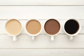 Different cups of coffee on white wooden table, top view. Gradient from light americano with milk to strong black espresso, copy space