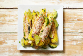 White plate of Mexican street corn on wooden background Mexican street corn shot straight down with room left and/or right for copy. Made with grilled corn with sour cream, cilantro sauce with grated parmesan and red pepper on top. Lime garnish to squeeze on top.