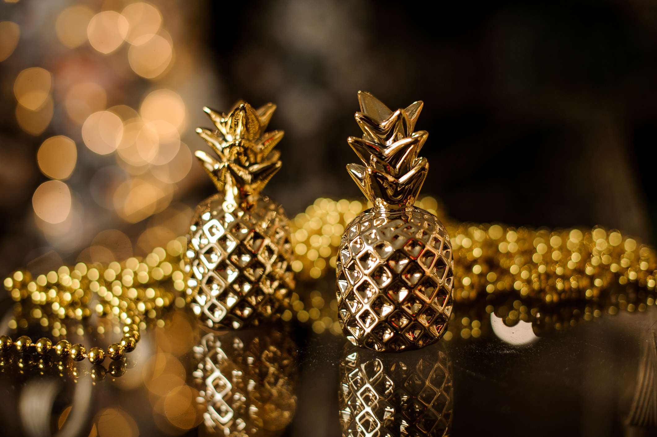 two gorgeous Christmas tree decorations in form of golden pineapples stand on glass table. Garland lights in background.