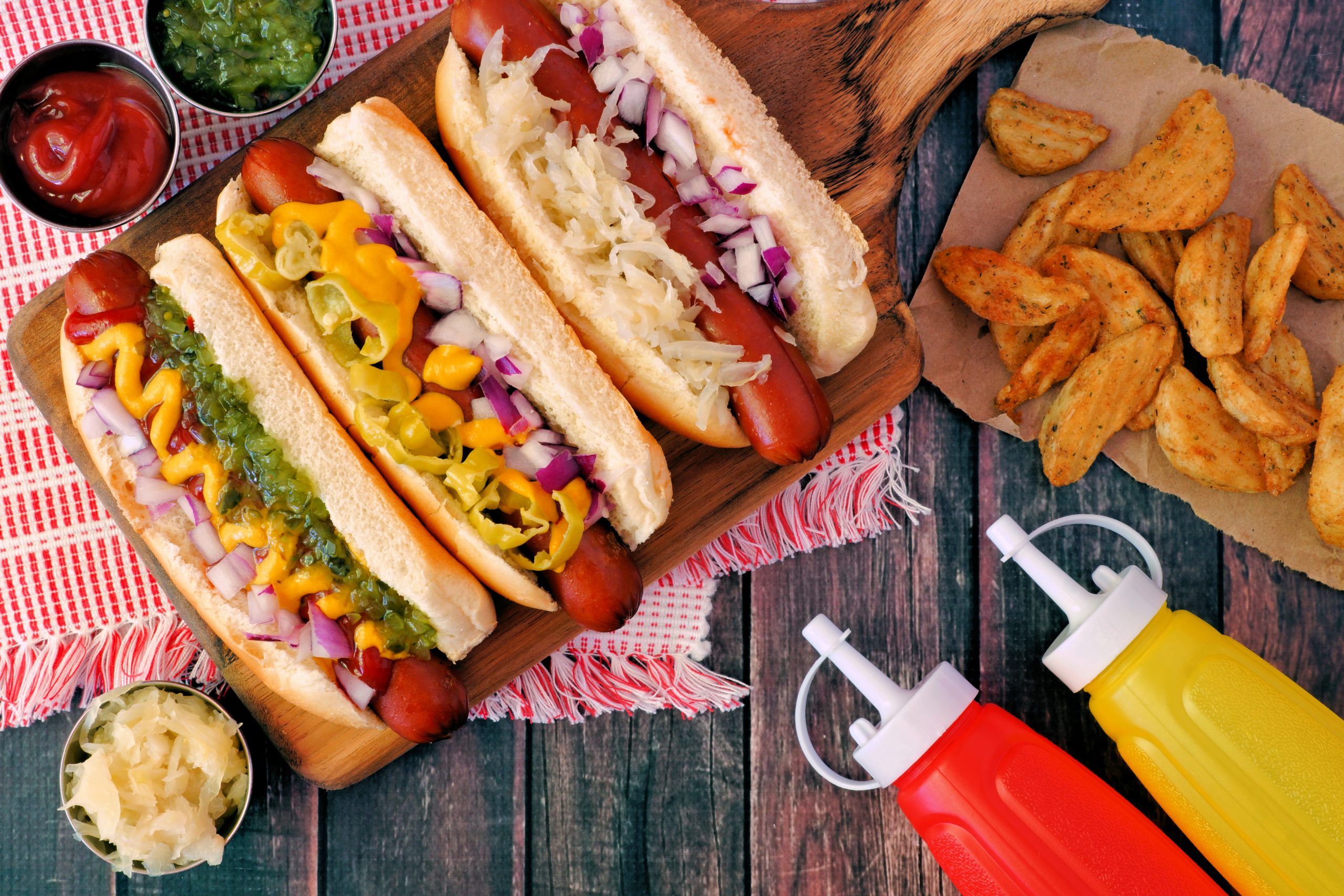 Hot dogs with assorted toppings and potato wedges, overhead scene on a rustic wood background