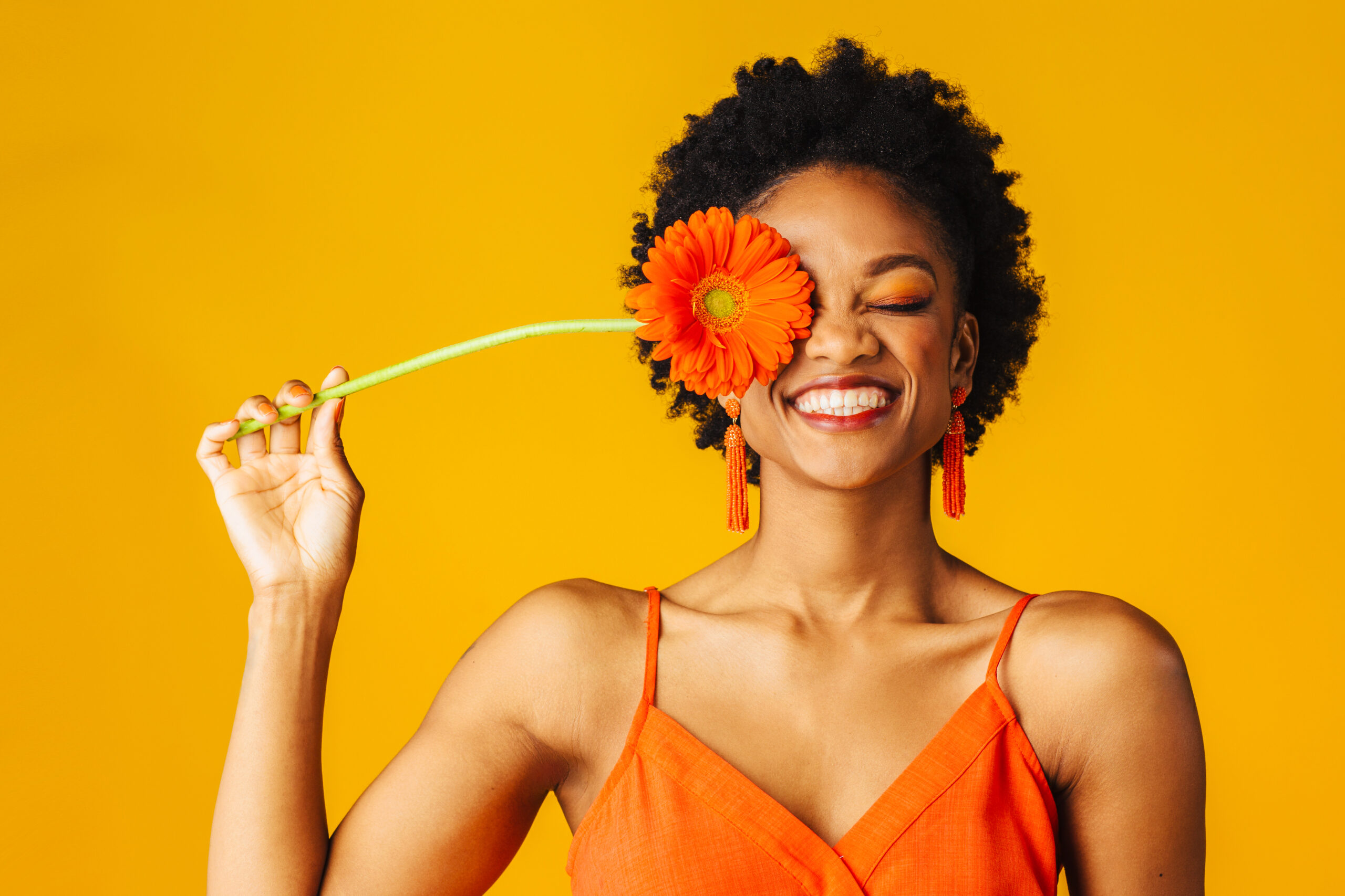 Portrait of a happy young woman holding orange Gerbera daisy covering her eye with eyes closed