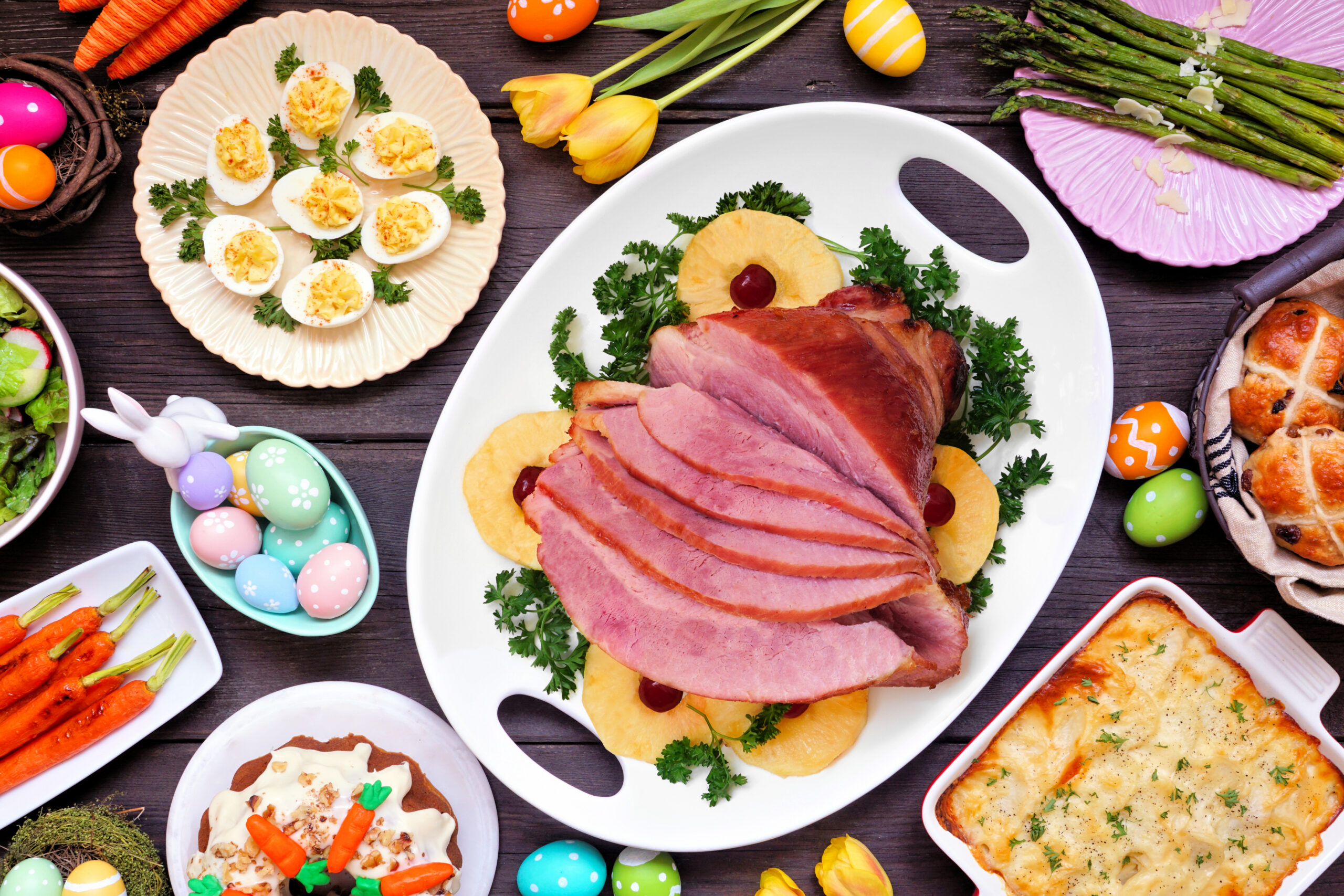 Traditional Easter ham dinner. Above view table scene on a dark wood background. Ham, scalloped potatoes, eggs, hot cross buns, carrot cake and vegetables.