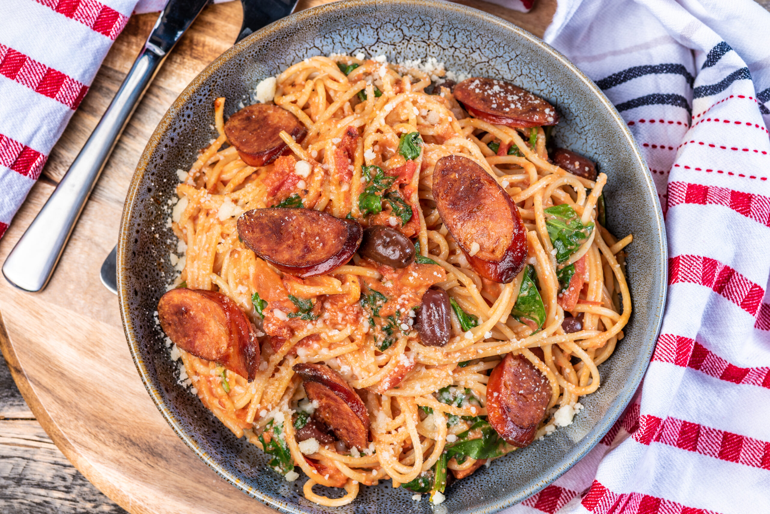 A bowl of chef prepared spaghetti with chorizo, black olives, and spinach in a creamy tomato sauce in a ceramic bowl on a wooden chopping board with red and white napery.