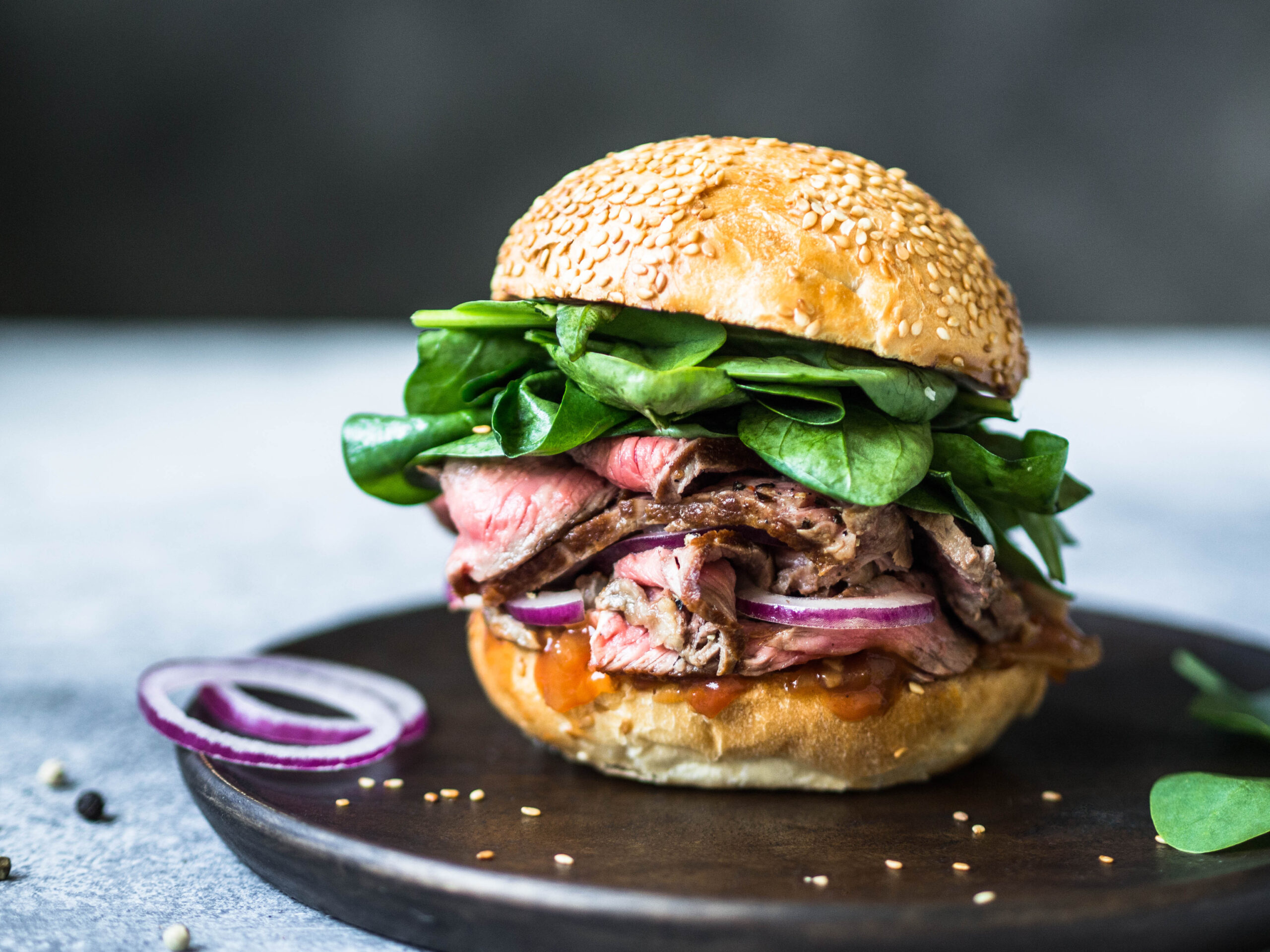 sandwich with steak slices, sauce of salsa, spinach and red onion. Burger from a bun with sesame and steak on a wooden plate on a gray background sandwich with steak slices, sauce of salsa, spinach and red onion. Burger from a bun with sesame and steak on a wooden plate on a gray background